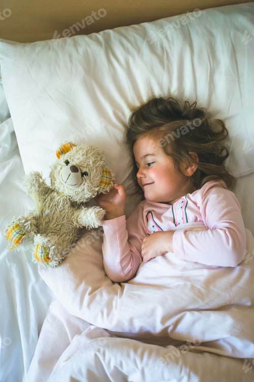 Preview: Little girl lying in a bed with teddy bear in the morning. Having fun at the beginning of the day