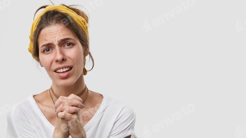 Preview: Concerned beautiful woman looks with upset facial expression, keeps hands in prayng gesture, wears h