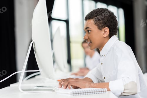 Preview: side view of african american teen boy typing on keyboard in front of computer screen in class