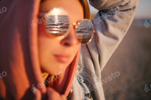 Preview: Young woman wearing casual clothes relaxing on a beach at sunset, ocean is reflecting in her glasses