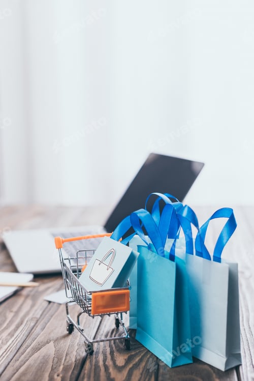 Preview: selective focus of toy shopping bags and shopping trolley with laptop on background