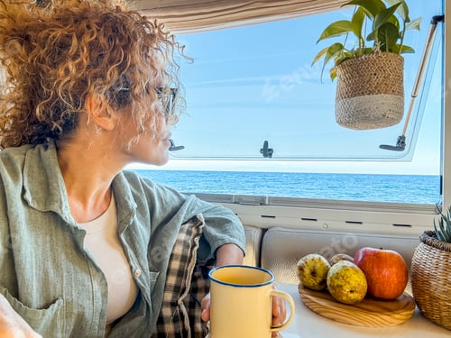 Preview: Curly woman with glasses smiles while drinking tea and looking out the window of her camper