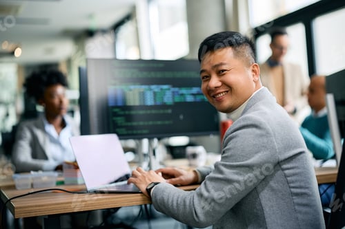 Preview: Happy Asian businessman working on laptop in the office and looking at camera.