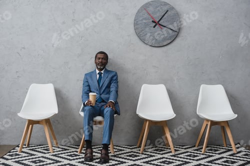 Preview: Handsome African american businessman sitting in a waiting room and holding coffee in a paper cup