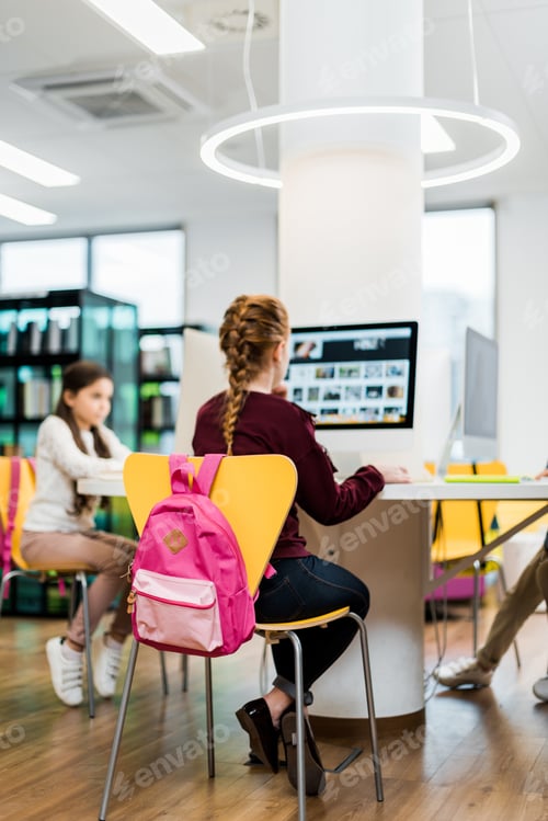 Preview: focused schoolgirls sitting and using desktop computers in library