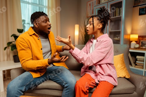 Preview: Young emotional African American couple, man and woman quarreling sitting in room at home conflict