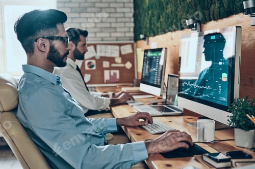 Preview: Two concentrated young modern men in formalwear working using computers while sitting in the office