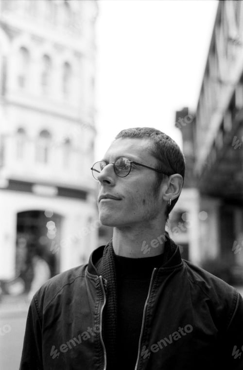 Preview: Young man wearing glasses and a black jacket in city street, monochrome analog photo