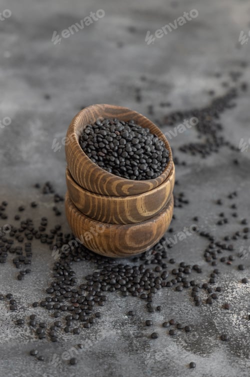 Preview: Wooden bowls of dry black lentils beans on grey table close up, protein source for vegetarian diet