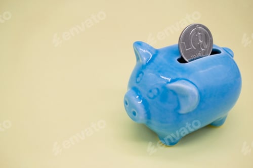 Vista previa: Blue ceramic piggy bank with a coin of 5 CZK in the hole on a yellow background. Copy space