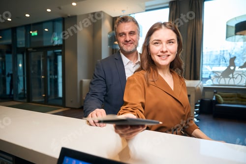 Preview: Woman giving passport at reception desk near her man standing in hotel lobby