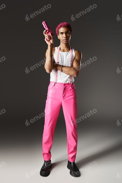 Preview: stylish african american man with pink hair pointing up his pink toy gun posing on gray backdrop
