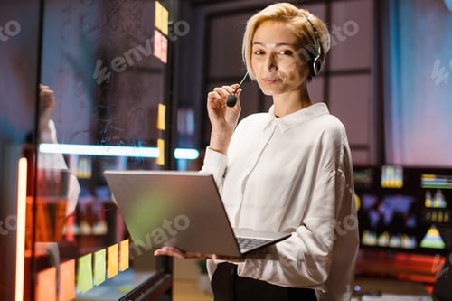 Preview: Caucasian businesswoman wearing headset using laptop, posing on camera near glass board at office