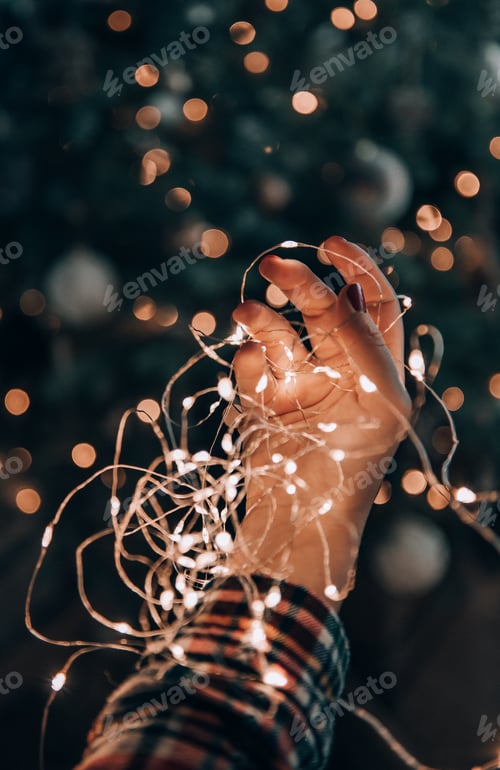 Preview: Close-up photo of female hand holding fairy lights in front of christmas tree