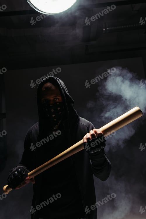 Preview: Silhouette of african american vandal with mask on face holding baseball bat on black background