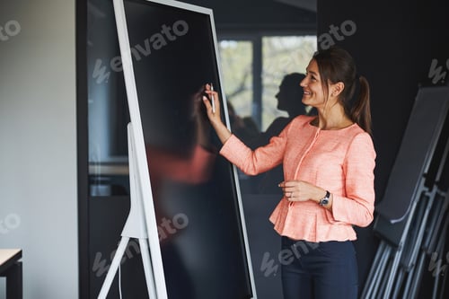 Preview: Happy attractive caucasian lady in pink jacket standing near the blackboard while writing something