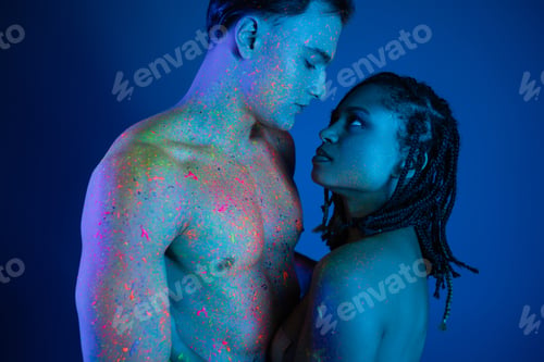 Preview: sexy multicultural couple in colorful neon body paint looking at each other on blue background with