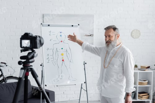 Preview: senior ayurveda coach pointing at human chakras system on flip chart in front of digital camera