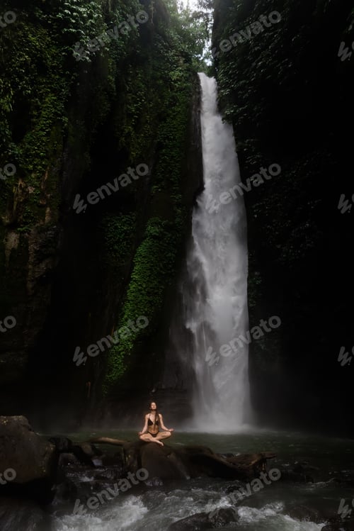 Preview: Beautiful young woman meditating while doing yoga in a wonderful forest near waterfall.