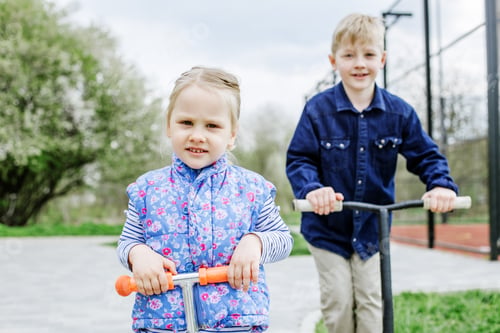 Preview: Little blond boy and girl ride kick scooters. Children play in playground on sunny spring day.