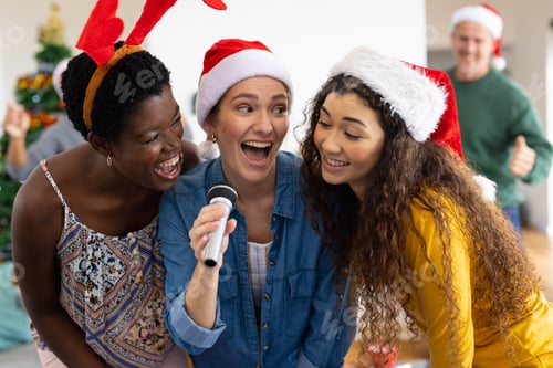 Preview: Image of happy diverse female friends celebrating christmas at home singing karaoke