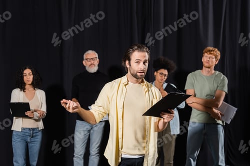 Preview: brunette bearded man holding clipboard and looking at camera while rehearsing near multicultural