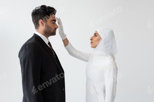Preview: side view of young bride in hijab touching forehead of groom during wedding ceremony isolated on