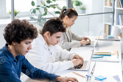 Preview: Group of three teenage students sitting in row by desk and working individually