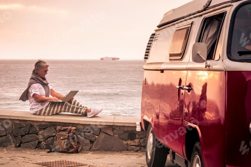 Preview: Senior woman travelling with a vintage van, sitting on wall by the sea, using laptop