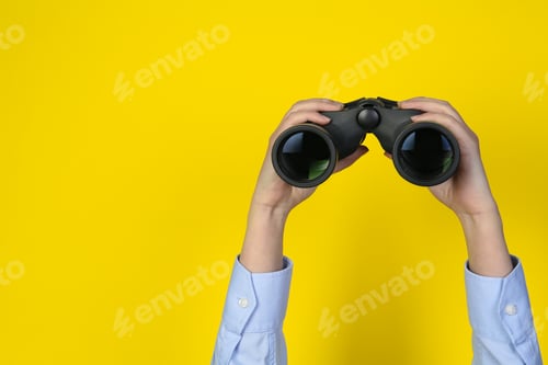 Preview: Woman holding modern binoculars on yellow background, closeup. Space for text