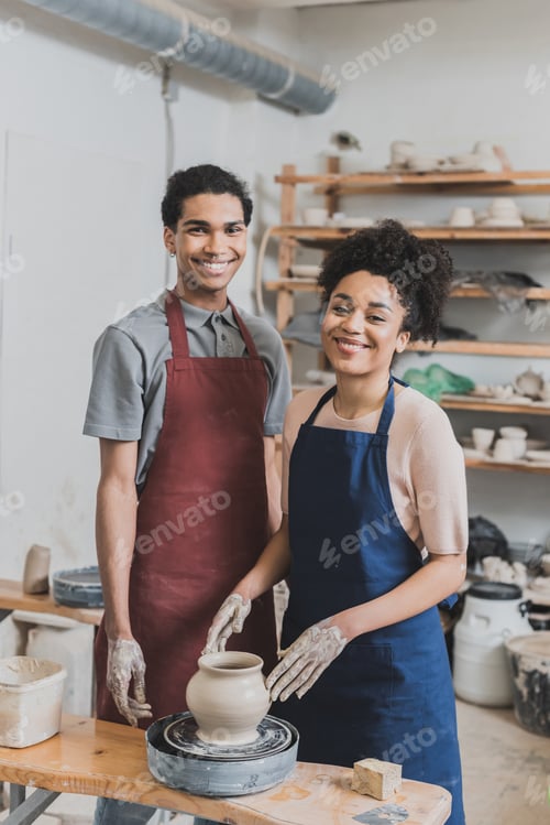 Preview: smiling young african american couple shaping wet clay pot on wheel in pottery