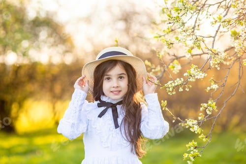 Preview: Portrait of cute little girl in a white dress and hat, standing under flowering trees, in spring