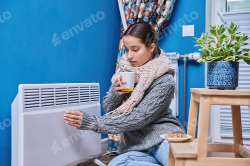 Preview: Teen female in sweater scarf warming near electric heating radiator, drinking hot drink