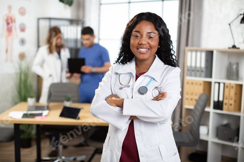 Preview: Afro-American doctor posing with arms crossed and diverse medical team working on the background