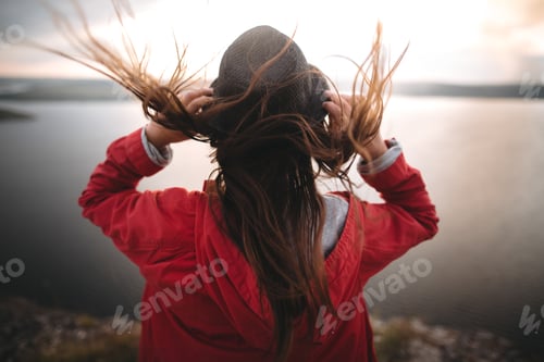 Preview: Girl in hat and with windy hair standing on top of rock mountain, enjoying beautiful sunset