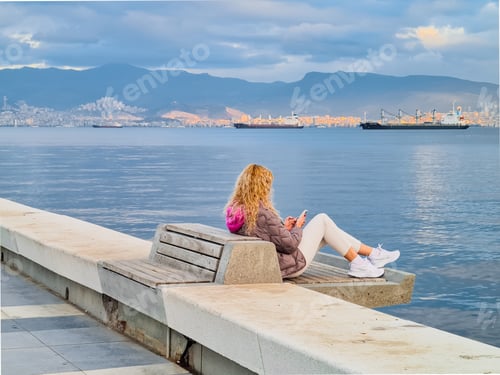 Preview: Blonde woman using her cellphone sitting on the bench on the sea embankment