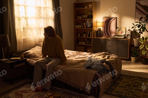 Preview: Young Woman Sitting on Bed Reflecting in Cozy Sunlit Bedroom