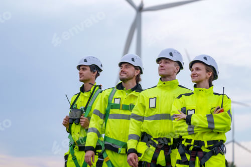Preview: Team of wind energy workers with safety gear stands near wind turbines at sunset