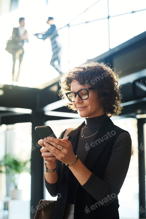 Preview: Young smiling female manager in eyeglasses watching video in smartphone