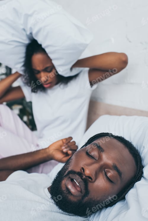 Preview: selective focus of african american woman covering head with pillow while sitting in bed near