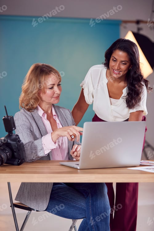 Preview: Mature Female Fashion Photographer With Client Looking At Images From Shoot On Laptop In Studio