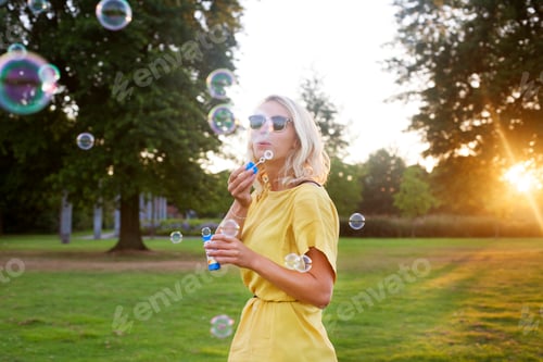 Preview: Portrait of young woman wearing yellow dress blowing bubbles in park at sunset