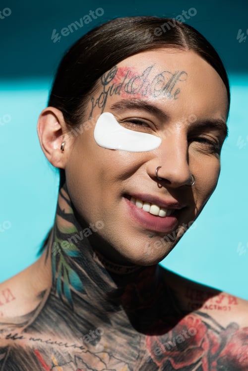 Preview: tattooed young man with eye patch smiling while looking at camera on turquoise