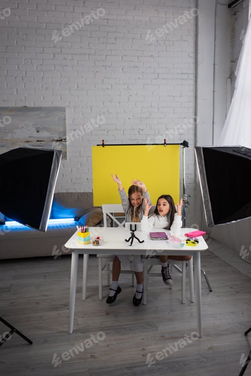 Preview: excited girls gesturing while recording video blog on mobile phone near lamps and yellow background