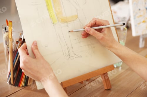 Preview: Woman drawing picture with pencil on easel at wooden table in artist's studio, closeup