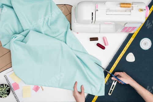 Preview: cropped shot of tailor cutting textile with scissors at workplace with various tailoring items