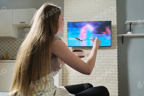 Preview: Young woman sitting at home watching tv and eating