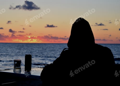 Preview: Male silhouette in a hood at a table in a cafe against the backdrop of the sunset sea