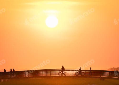 Preview: people in silhouette riding bikes on the Long Beach boardwalk at sunset with an orange glow