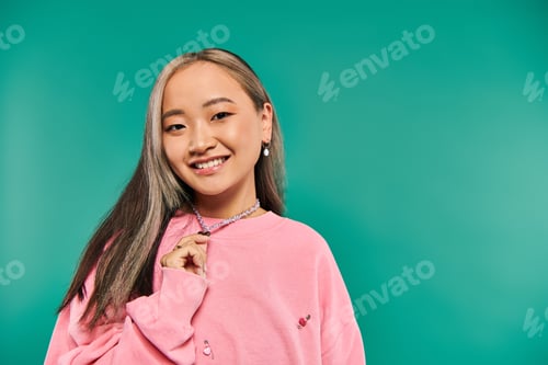 Preview: portrait of cheerful asian woman in pink sweatshirt pulling heart shaped necklace on blue backdrop